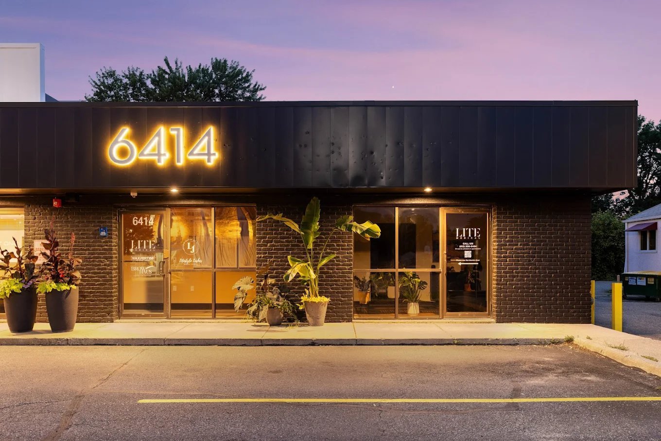 Storefront with brick facade and illuminated golden neon number 6414 sign at dusk, with plants flanking entrance doors